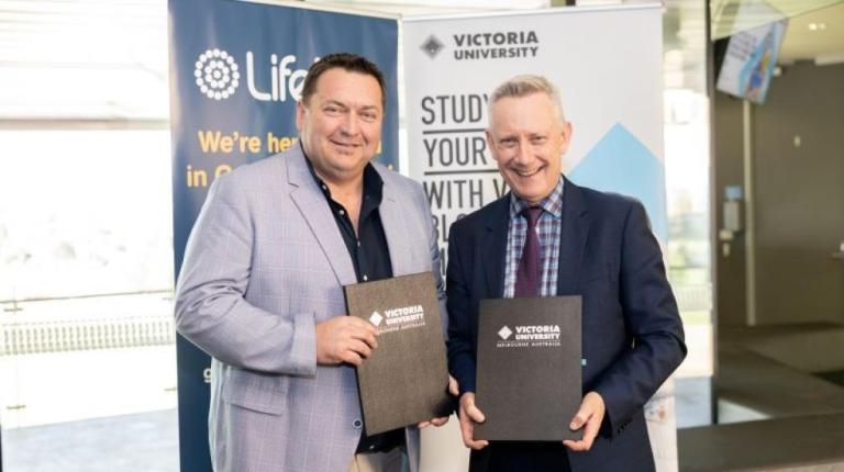  Two men in suits hold paperwork in front of Lifeline and Victoria University signs
