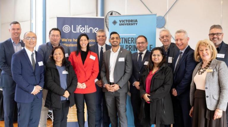  A group of men and women in work clothes pose in front of Victoria University and Lifeline banners