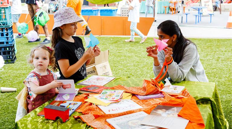 Toddler and child fold paper at an outdoor table, with the help of an adult