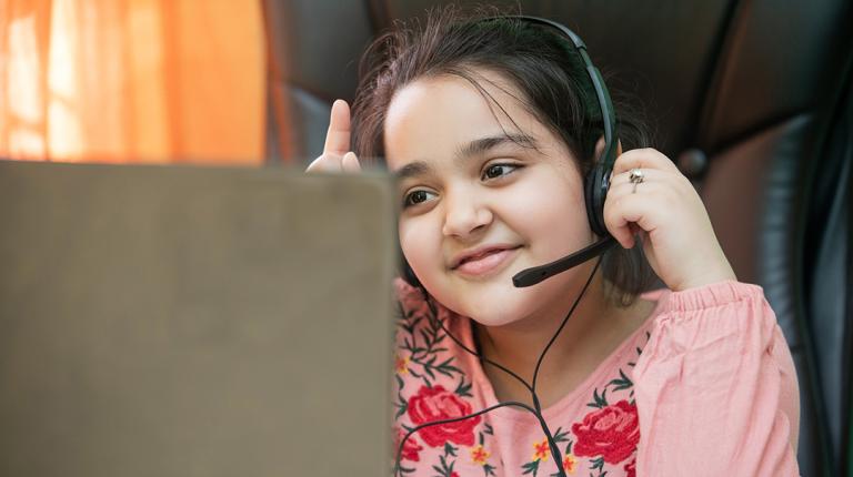 a school-aged girl with a headset sits in front of a computer with a hand raised