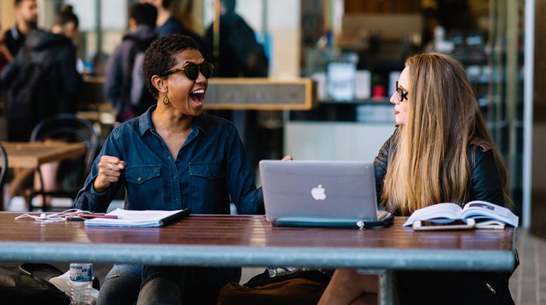  Two students sit outside at a table laughing together. 