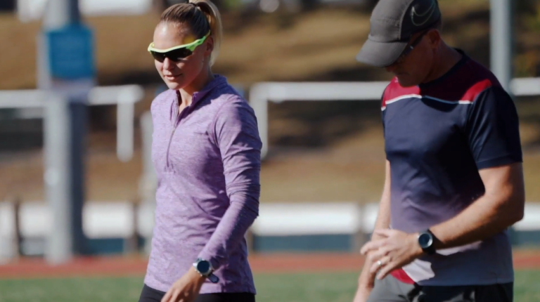 Woman on sport field in sport attire wearing dark glasses, with man in sport attire