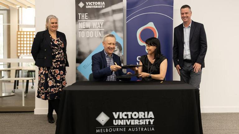 A man and woman smile at a Victoria University branded table, with branded banners in the background and two smiling, professional looking people