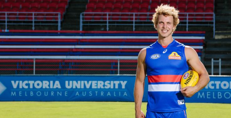 Mitch Wallis in his Bulldogs gear stands in front of a VU banner