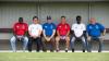 Group of 6 men from diverse ethnic backgrounds smile at the camera from a footy bench, wearing red white and blue
